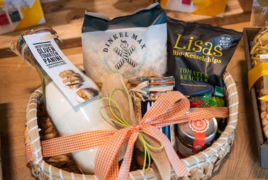 Geschenkkorb mit verschiedenen Lebensmitteln, darunter Mehl, Kekse und Chips, dekoriert mit einem orange-weiß karierten Band