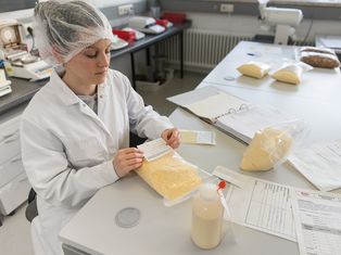 Mitarbeiterin der Qualitätssicherung in weißem Kittel und Haarnetz prüft Mehlproben in einem Labor mit mehreren Mehlpackungen und Dokumenten auf dem Tisch