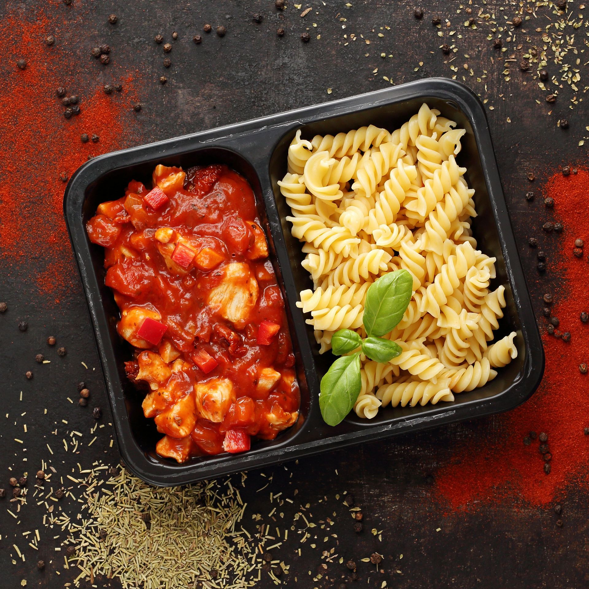 Schwarze Plastikschale mit Pasta und Tomatensauce, garniert mit Basilikum, umgeben von Gewürzpulvern