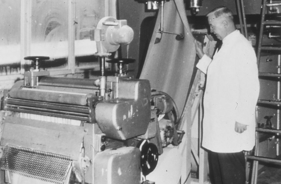 A man in a white coat stands next to a pasta machine, it is a black and white picture