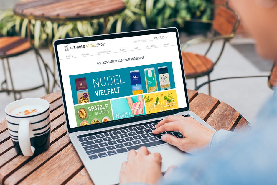 Laptop zeigt eine Webseite mit dem Titel 'Nudel Vielfalt' und verschiedenen Nudelbildern, Person bedient das Touchpad an einem Holztisch mit Kaffeetasse