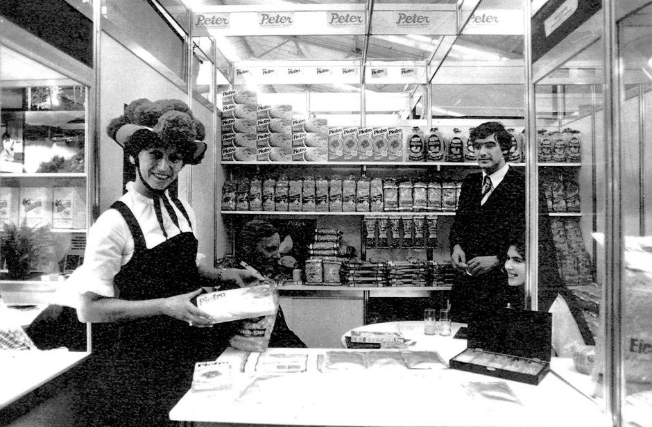 Two people in a grocery store, a woman with an apron holding a tray, a man standing behind the counter with shelves full of products