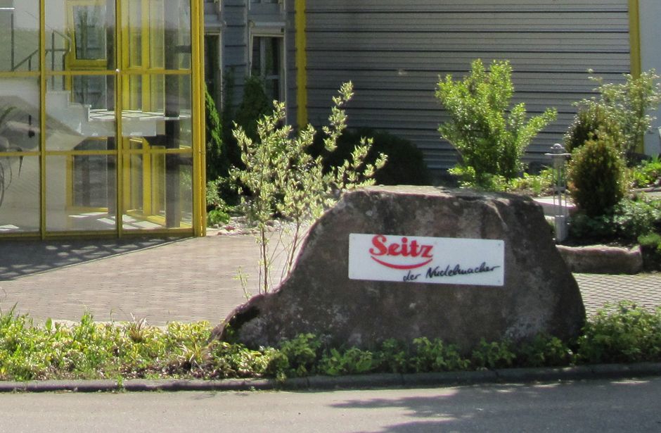 Large stone with sign ‘Seitz’ in front of a building with glass facade and plants