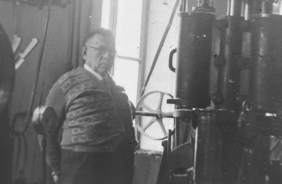 Elderly man standing next to a large industrial machine in a workshop in black and white