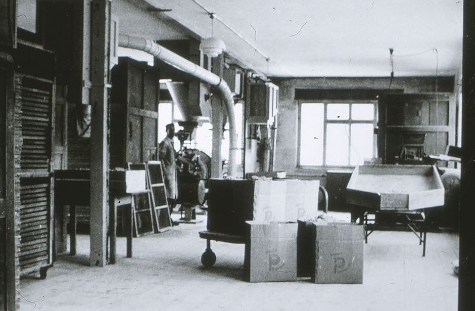 Black and white image of a factory hall with machines, pipes and cardboard boxes on the floor
